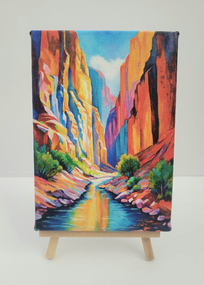 A colorful canvas painting of a canyon landscape with a river, displayed on a small wooden easel against a plain light background.