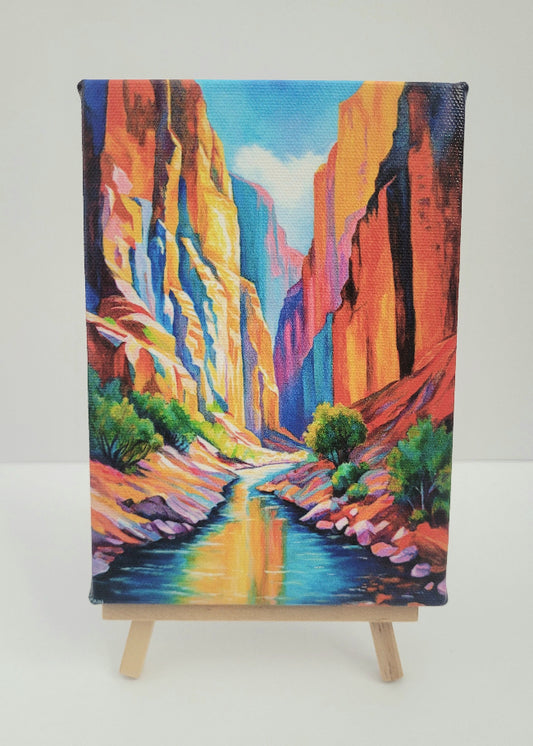 A colorful canvas painting of a canyon landscape with a river, displayed on a small wooden easel against a plain light background.