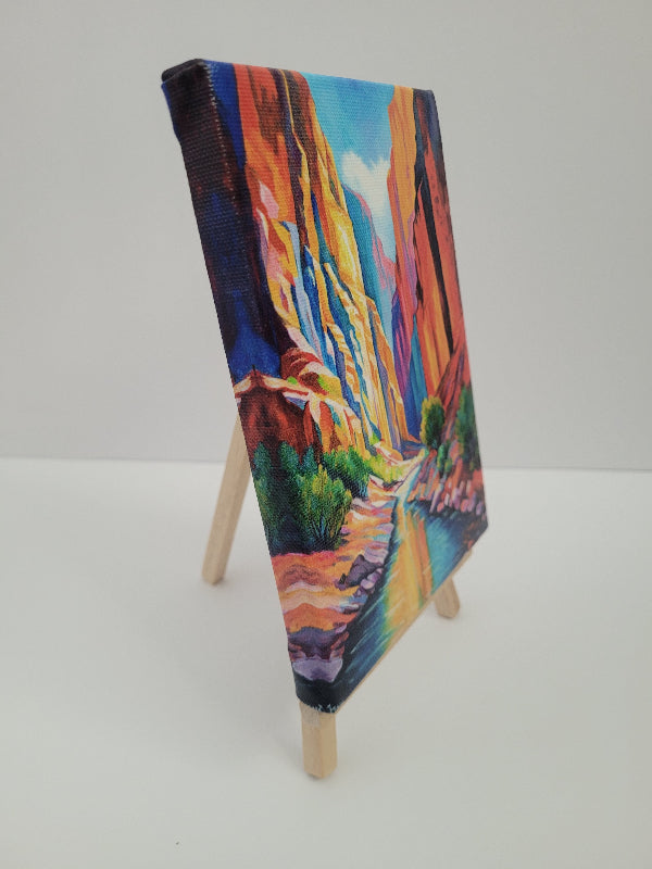 A small canvas painting of a colorful canyon landscape with a river, displayed on a miniature wooden easel against a plain white background.